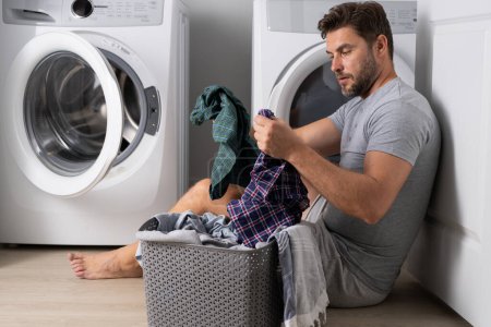 Téléchargez les photos : Homme faisant la lessive à la maison près de la machine. Un mari qui fait la lessive à la maison. Sexy gars laveuse mettre des vêtements dans la machine à laver. Concept de ménage. Laveuse et sécheuse, laverie automatique - en image libre de droit