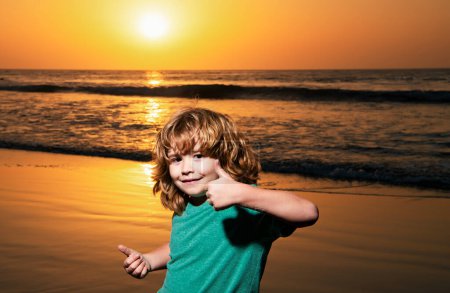 Téléchargez les photos : Petit garçon drôle sur la plage du coucher du soleil, pouce levé. Incroyable surpris enfants émotions - en image libre de droit
