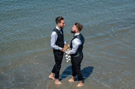Schwule Bräutigame spazieren während des Hochzeitstages gemeinsam am Strand. Romantische Männer im Meerwasser. Romantisches und sinnliches homosexuelles Paar. Schwule Bräutigame am Hochzeitstag zusammen