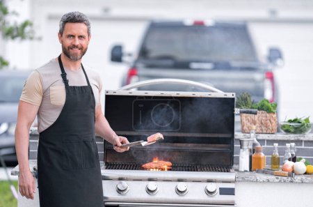 Téléchargez les photos : Portrait d'un jeune homme joyeux griller de la viande sur le barbecue en plein air.. Grillage et grillage des aliments. Faire rôtir la viande à l'extérieur. Barbecue et grill. Cuisson de viande dans la cour arrière - en image libre de droit