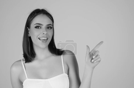 Photo for Young happy woman pointing copy space. Studio close up portrait of Girl looking at camera and pointing away on isolated background. Smiling Girl points a finger away. Young woman showing copy space - Royalty Free Image