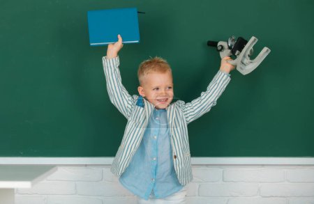 Photo for Portrait of funny pupil of primary school study in classroom at school. Little child with microscope and book - Royalty Free Image