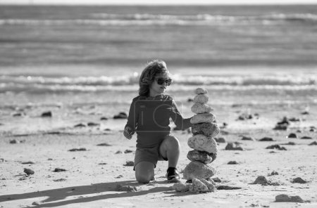 Téléchargez les photos : Méditation estivale pour enfants. Enfant avec des pierres zen sur la plage de la mer, méditation, spa et harmonie. Calme concept d'équilibre. Les enfants jouent sur la plage. Calmant enfant et tour de pierre contre la mer - en image libre de droit