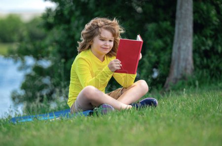 Téléchargez les photos : Smart enfant garçon livre de lecture dans le parc en plein air. Un gamin blond lisant un livre en été. Portrait extérieur intelligent pour enfants. Enfants imagination, innovation et inspiration. École en plein air, apprendre les enfants - en image libre de droit