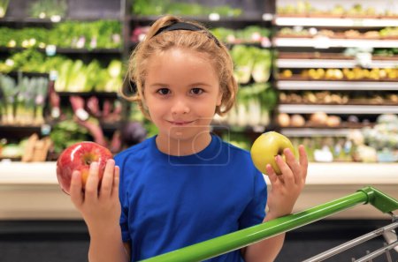 Foto de Niño con manzana y carrito de compras en el supermercado. Compras en el supermercado. Niños comprando comestibles en el supermercado. El niño compra verduras frescas en la tienda de comestibles. Niño en la tienda - Imagen libre de derechos