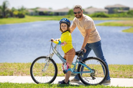 Foto de Feliz día de los padres. Padre enseñando a su hijo a montar bicicleta. Padre ayudando a su hijo emocionado a montar en bicicleta en el vecindario americano. Niño en casco de bicicleta aprendiendo a andar en bicicleta con su padre. Día de los padres - Imagen libre de derechos