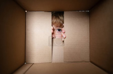 Téléchargez les photos : Joyeux petit garçon ouvre cadeau et regarder à l'intérieur de la boîte en carton. Fermez les yeux en regardant - en image libre de droit