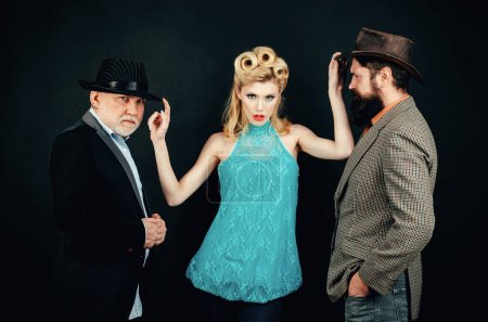 Two men in suits and a girl in an evening dress. Funny gangsters in hat with young woman. Polyamorous relationship. Modern partnership. Confident woman dominating over two men. Old and young