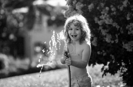 Foto de Niño adorable regando las plantas, desde el spray de manguera con manguera de agua en el jardín en el patio trasero de la casa en una noche de verano - Imagen libre de derechos