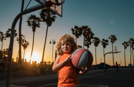 Téléchargez les photos : Jeu de basket-ball pour enfants. Mignon petit garçon tenant une balle de basket-ball essayer de faire un score. Adorable enfant jouant au basket au tribunal au coucher du soleil - en image libre de droit