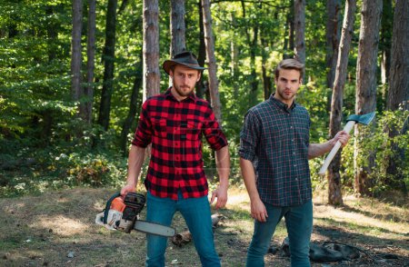 Woodcutters with chainsaw lumberjacks with axe. Hipsters men on serious face with axe. Lumberjack brutal and bearded holds axe. Two lumberjacks in forest. Forest workers