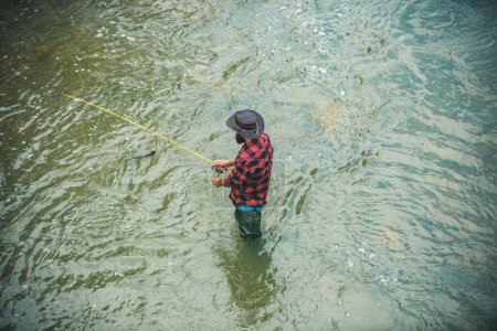 Téléchargez les photos : Jeune homme barbu pêchant dans un lac ou une rivière. Pêche à la mouche. Pêcheur homme dans l'eau attraper du poisson, vue de dessus - en image libre de droit