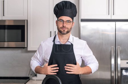 Foto de Hombre guapo chef en uniforme de cocina en la cocina. Concepto de menú del restaurante. Hombre hispano con uniforme de panadero. Cocina y gastronomía. Chef masculino en uniforme de trabajo, delantal negro, sombrero de chef - Imagen libre de derechos