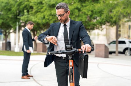 Téléchargez les photos : Homme d'affaires attrayant en scooter de pied au parc de New York. Homme d'affaires en costume sur un scooter électrique lors d'une réunion d'affaires. Homme d'affaires d'âge moyen équitation scooter en costume - en image libre de droit