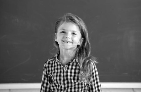 Photo for Close-up portrait of attractive small little cheerful girl sitting on table desktop in class room indoors. Little funny school girl face. Kids education and knowledge. Student kids - Royalty Free Image