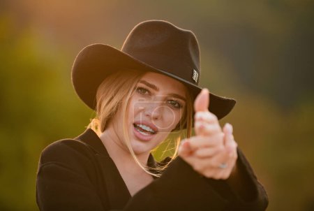 Téléchargez les photos : Belle jeune femme simule le tir avec les doigts. Cowgirl en chapeau de cow-boy avec pistolet à doigt. Mode et beauté - en image libre de droit