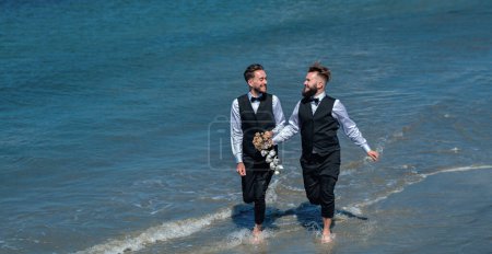 Foto de Los novios gay caminando juntos en la playa del mar durante el día de la boda. Hombres románticos en el agua de mar. Feliz pareja gay en la boda. Pareja gay romántica y sensual. gay novios juntos en boda día - Imagen libre de derechos