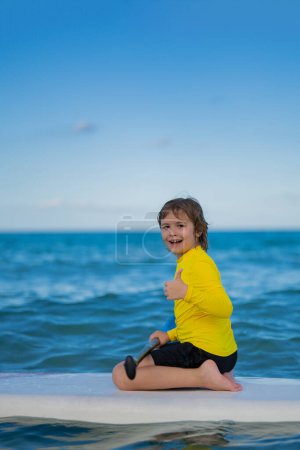 Kid SUP boarder. Kind beim Paddeln auf dem Paddelbrett oder auf dem Sofa. Gesunder Lebensstil im Sommer. Sommer-Wassersport. SUP-Surftour im Sommer am Strand. Junge schwimmt mit Paddel und Brett