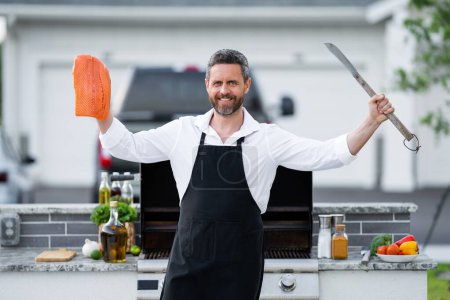 Photo for Man in chef apron hold salmon fish at barbecue grill. Male cook preparing barbecue outdoors. Bbq fish, grill for picnic. Roasted salmon fish. Grilled fish and barbecue seafood concept - Royalty Free Image