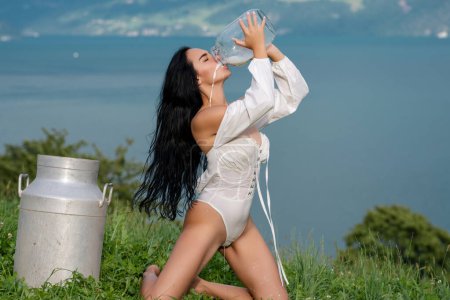 Foto de Country Girl con una botella de leche fresca en el pueblo. Suiza. Mujer joven en lencería bebiendo leche. Mujer joven bebiendo leche de la botella. Belleza chica romántica beber leche al aire libre - Imagen libre de derechos