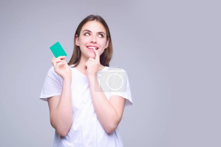 Foto de Mujer joven en camiseta blanca con tarjeta de crédito cerca de la cara, aislada sobre fondo gris. Paga con tarjeta de crédito o débito. Tarjeta bancaria. Tarjeta de crédito de plástico - Imagen libre de derechos