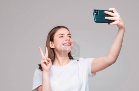 Foto de Emociones felices, chica sobre fondo gris tomando selfie en el teléfono móvil, el uso de la aplicación de fotos y mostrar signo de paz cerca de la cara y sonriendo - Imagen libre de derechos