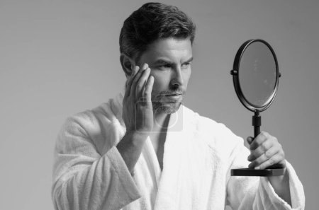 Man applying cosmetic face cream on mirror. Facial treatment. Portrait of man with bare naked shoulders touching skin. Sexy man with skin care product. Male face creme. Perfect skin, morning routine