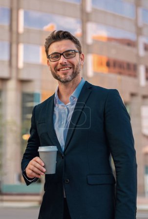 Foto de Hombre de negocios alegre en vasos con café para ir caminando por la calle hacia el moderno edificio de oficinas. Hombre de negocios hispano sonriendo feliz en la ciudad. Retrato de hombre de negocios al aire libre - Imagen libre de derechos