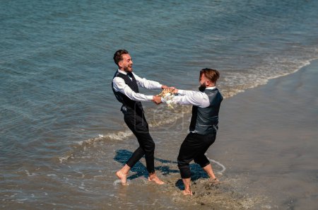 Photo for Romantic men same sex marriage. Gay grooms walking together on sea beach during Wedding day. Romantic men in sea water - Royalty Free Image