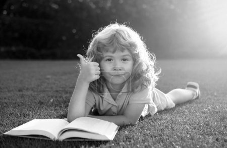 Téléchargez les photos : Joli garçon avec les pouces vers le haut livre de lecture sur l'herbe verte. Été plein air enfants éducation - en image libre de droit