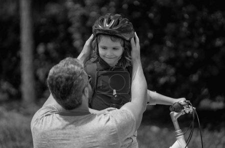 Father and son riding bike in park. Child in safety helmet with father riding bike in summer day. Father teaching son riding bike. Fatherhood. Father helping his son to wear a cycling helmet