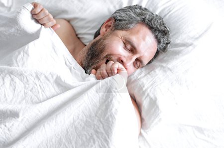 Foto de Cabeza masculina sobre almohada. Hombre canoso bostezando en la cama blanca. Hombre mayor bostezando en la cama en el dormitorio. Un hombre maduro bostezando en casa por la mañana. Buen sueño. Millennial hombre bostezando en la cama - Imagen libre de derechos