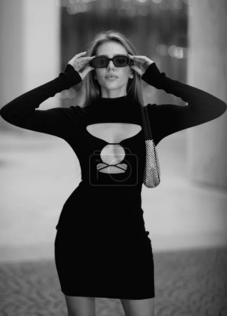 Téléchargez les photos : Modèle de mode en lunettes de soleil de mode, robe sexy noire et sac à main dans la rue. Fille à la mode en vêtements à la mode posant dans la rue de la ville. Portrait style de vie Vogue - en image libre de droit