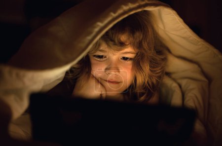 Photo for Kid lying in bed at night and playing digital tablet. Child watching tablet. Child in bed at night watching tablet, face illuminated by light of screen. Kid with tablet on bed watching cartoons - Royalty Free Image