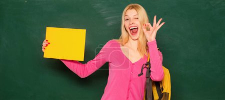 Photo for Excited amazed teacher. Student girl holding empty faper for product. Portrait of smiling female teacher near blackboard in classroom at school - Royalty Free Image