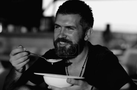 Foto de El hombre come sopa. Buen apetito. Hombre barbudo con un tazón de sopa. Un tipo feliz comiendo sopa al aire libre. Hombre comiendo deliciosa sopa en el restaurante. Cena y comida. Hombre almorzando en un café - Imagen libre de derechos