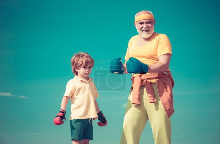 Photo for Portrait of a determined senior boxer with little boy child over blue sky background. Boxer grandfather and child with blue boxing glove on blue sky background - isolated - Royalty Free Image