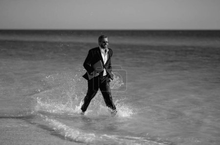 Téléchargez les photos : Homme d'affaires costume pieds nus courir sur la plage. Beau homme d'affaires en vacances d'été. Homme d'affaires en cours d'exécution sur la mer tenant ordinateur portable. Homme d'affaires excité sauter dans l'eau de mer. Homme d'affaires fou - en image libre de droit