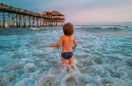 Foto de Niño jugando al aire libre saltando al agua en las vacaciones de verano en la isla de playa tropical. Feliz niño jugando en el mar. Océano azul con grandes wawes blancos en el fondo - Imagen libre de derechos