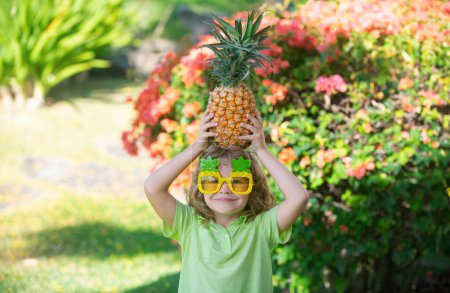 Téléchargez les photos : Joyeux petit enfant tient un ananas sur la tête sur un fond de nature estivale - en image libre de droit