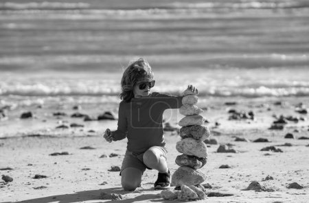 Photo for Summer kid meditation. Child play with pyramid stones balance on the sand of the beach. Kid with stack of stones on sand near sea. Child play with stones balance on beach. Balance and harmony concept - Royalty Free Image