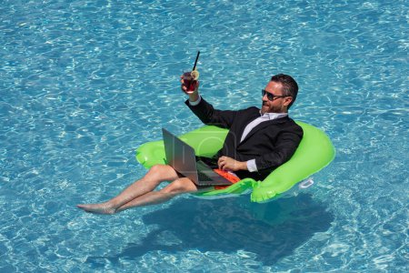 Téléchargez les photos : Homme d'affaires prospère flottant en costume dans l'eau de la piscine. Vacances d'été et concept de voyage. Homme d'affaires drôle boire cocktail d'été et en utilisant un ordinateur portable dans la piscine. Concept d'entreprise d'été - en image libre de droit