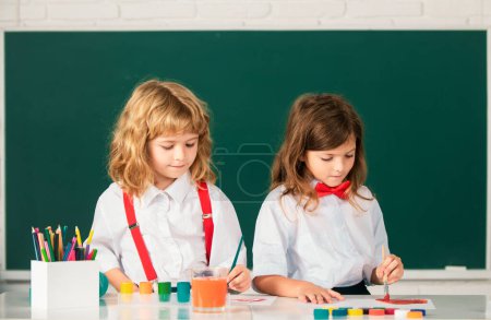 Photo for School children girl and boy painting with paints color and brush in classroom. Siblings drawing with pencil and paints - Royalty Free Image