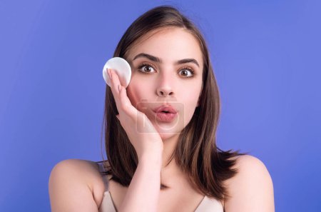 Foto de Beautiful young woman with cotton pad, applying lotion on face, removing makeup. Skin care. Studio portrait of girl cleaning her face with cotton pad, isolated background. Face toner and cotton pad - Imagen libre de derechos