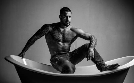 Sexual macho man in bath. Strong muscular tattoed man holding champagne bottle and posing in bathroom. Handsome bearded shirtless man in jeans with sexy body in bathroom