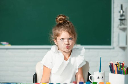 Téléchargez les photos : Enfant fille dessine dans la salle de classe assis à une table, s'amusant sur fond de tableau noir de l'école. drôle école fille visage - en image libre de droit