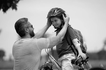 Téléchargez les photos : Petit enfant apprenant à faire du vélo avec son père dans le parc. Père enseignant le cyclisme fils. Père et fils apprennent à faire du vélo à la fête des Pères. Père aidant son fils à porter un casque de cyclisme - en image libre de droit