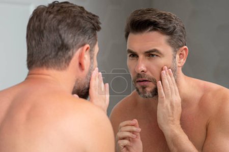 Téléchargez les photos : Homme hispanique d'âge moyen regardant dans le miroir, la peau du visage et le chaume. Produit de soins de beauté masculin. Soins de la peau, spa à domicile. Portrait de beauté d'un bel homme. Modèle spa, crème nourrissante hydratante - en image libre de droit