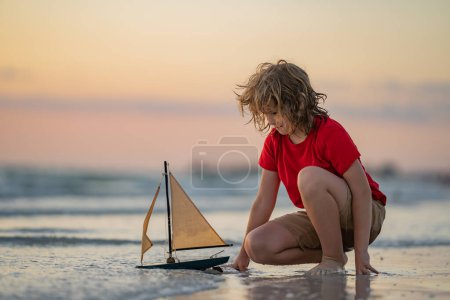 Téléchargez les photos : Enfant avec bateau jouet dans l'eau de mer en vacances d'été. Petit garçon jouant avec jouet bateau à voile sur la mer. Vacances d'été, garçon enfant jouant avec le bateau jouet à la plage. Vacances avec les enfants. Enfant avec bateau jouet - en image libre de droit