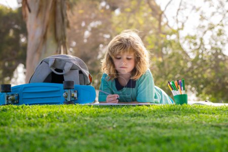 Foto de Niño con tarea de arte en el patio de recreo. Niño lindo con escritura a lápiz en el cuaderno al aire libre. Concepto de aprendizaje y educación para niños al aire libre. Tarea de vacaciones de verano - Imagen libre de derechos